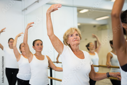 Group of ballerina standing with one hand on barre and reaching over hand in bright fitness room