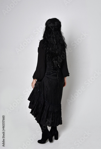 Full length portrait of dark haired woman wearing black victorian witch costume. standing pose, back view, with gestural hand movements, against studio background.