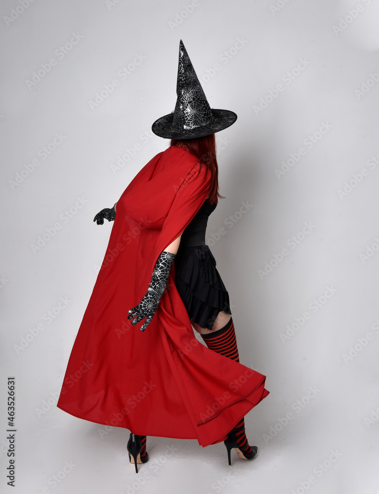 Obraz premium Full length portrait of dark haired woman wearing black victorian witch costume with a flowing cloak. standing pose, with gestural hand movements, against studio background.