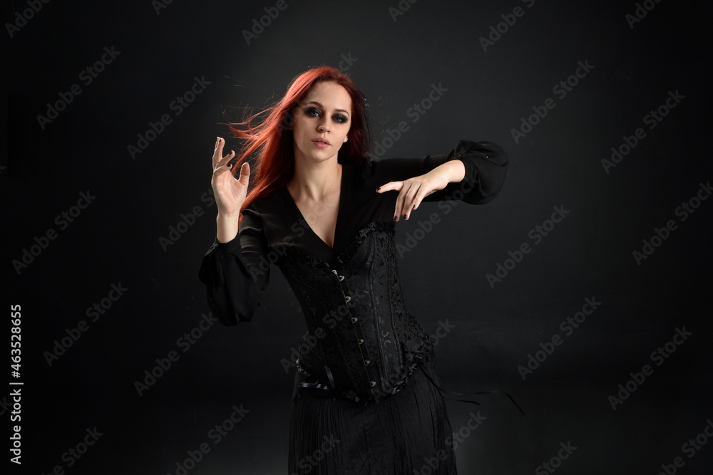 close up portrait of red haired woman wearing  black victorian witch costume.  standing pose, with  gestural hand movements,  against dark studio background.