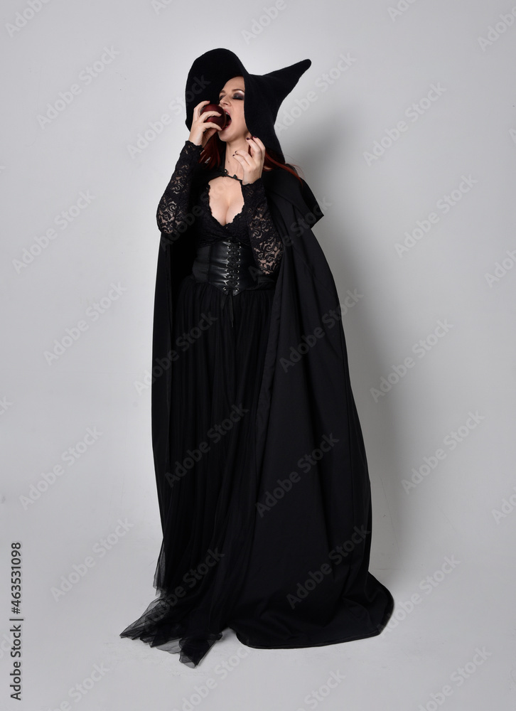 Full length portrait of dark haired woman wearing  black victorian witch costume  standing pose, with  gestural hand movements,  against studio background.