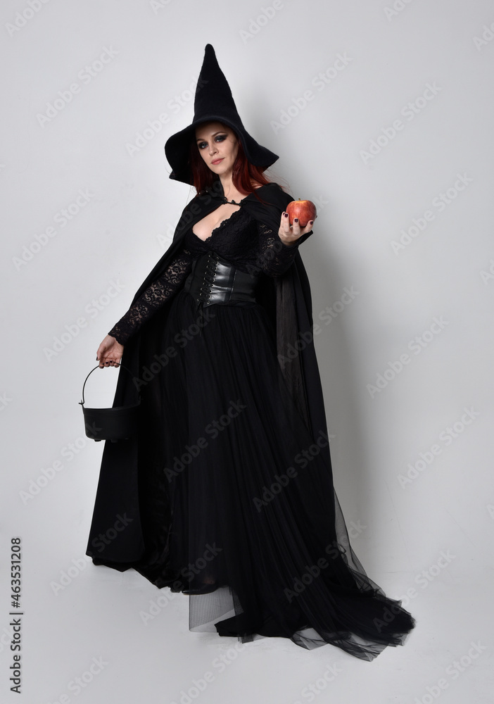 Full length portrait of dark haired woman wearing black victorian witch costume standing pose, with gestural hand movements, against studio background. Full length portrait of dark haired woman wearing black victorian witch costume standing pose, with gestural hand movements, against studio background.
