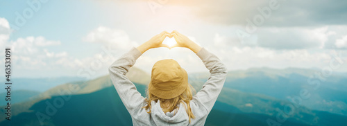 Back view of adventure traveler female in hat standing over summer mountain peaks and making shape of love heart by hands. Travel and explore concept © Shi 