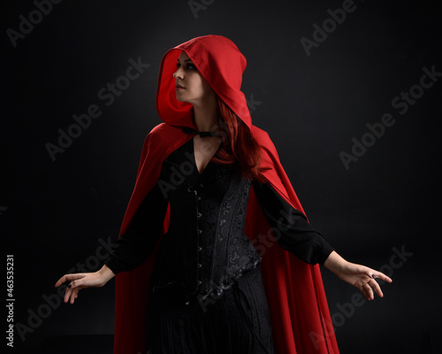 close up portrait of girl wearing red riding hood cloak and witch costume, isolated on black studio background with shadowy rim lighting. 