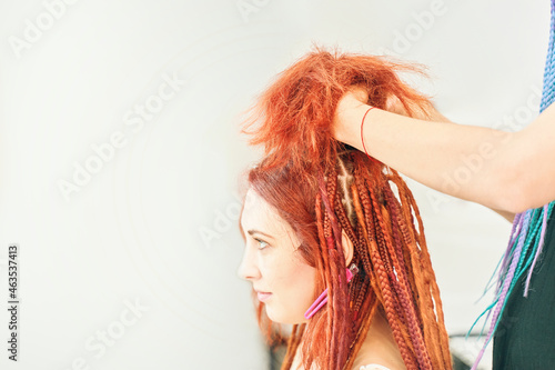 Process of braiding braids on head in beauty salon close up. Red-haired girl makes braids-dreadlocks. Dreadlocks from kanekalon. Cool fashion hairstyle made of artificial hair. Barber services.