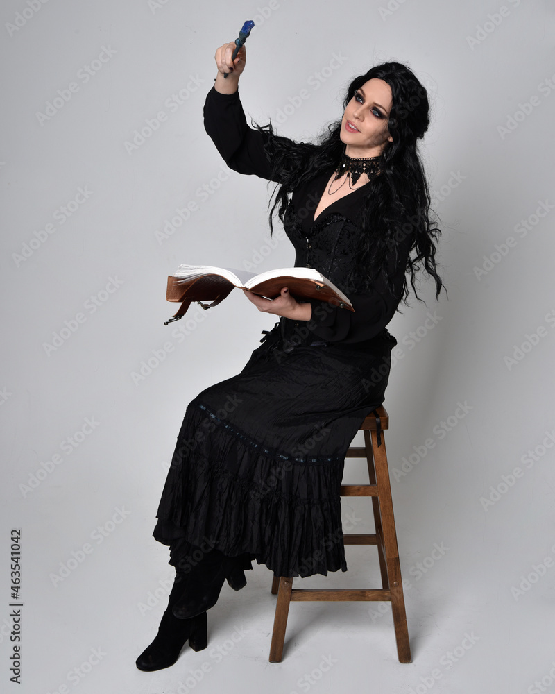 Full length portrait of dark haired woman wearing  black victorian witch costume.  sitting pose on a chair, with gestural hand movements,  against studio background.