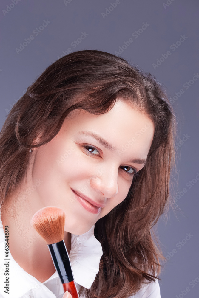 Makeup Concepts. Beautiful Caucasian Young Woman Posing with Make-up Brush Near Face and Smiling Over Gray Background. Makeup Concepts. Beautiful Caucasian Young Woman Posing with Make-up Brush Near Face and Smiling Over Gray Background.