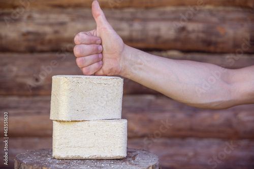 Fuel briquettes from pressed sawdust against the background of a log wall. Hand with a raised thumb. photo