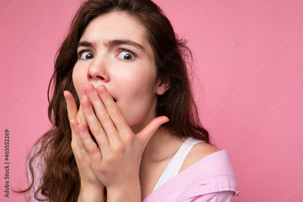 Closeup of young shocked astonished beautiful curly brunet female person with sincere emotions wearing casual pink shirt isolated over pink background with free space and covering mouth with hands Closeup of young shocked astonished beautiful curly brunet female person with sincere emotions wearing casual pink shirt isolated over pink background with free space and covering mouth with hands