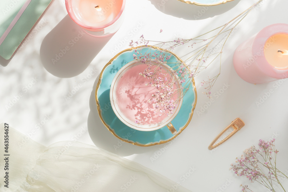 Pink flower tea in vintage porcelain tea cup, candles, flowers and notebook on white table with natural light. Romantic morning routine with healthy warm drink. Top view. - obrazy, fototapety, plakaty 