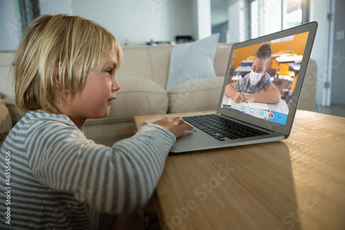 Caucasian boy using laptop for video call, with elementary school pupil with face mask on screen