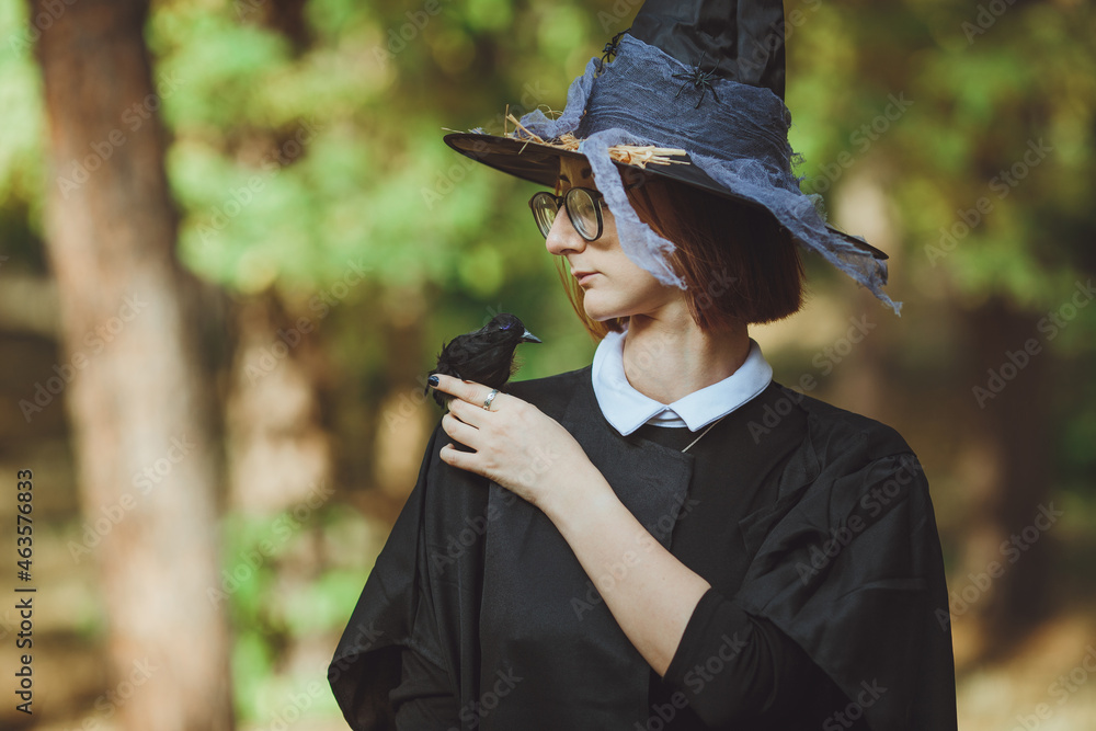 Halloween witch. Portrait of a girl in a hat and witch costume and with a black raven in the forest. Ready for the holiday wallet or life