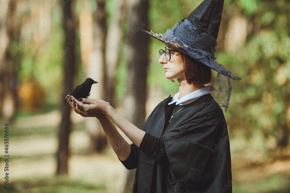 Halloween witch. Portrait of a girl in a hat and witch costume and with a black raven in the forest. Ready for the holiday wallet or life