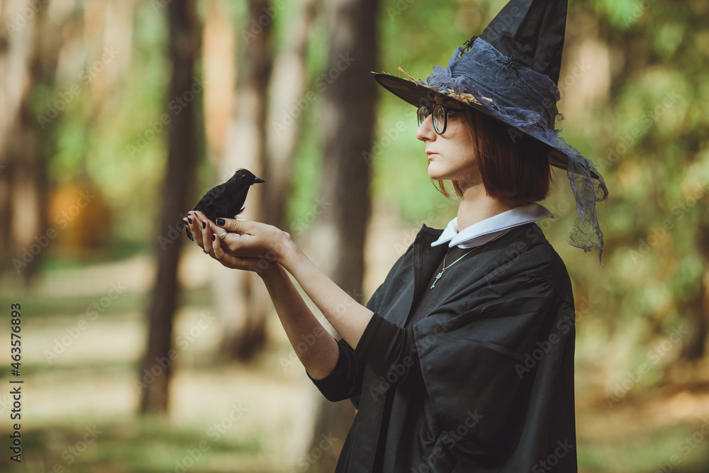 Halloween witch. Portrait of a girl in a hat and witch costume and with a black raven in the forest. Ready for the holiday wallet or life