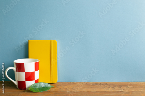 Obraz na płótnie Yellow diary note book with mug cup on wooden desk. blue wall background w salonie