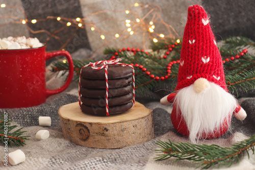 Funny Christmas card with toy gnome, a cup of hot chocolate with marshmallows, chocolate coockies on wooden cut and fir tree branches