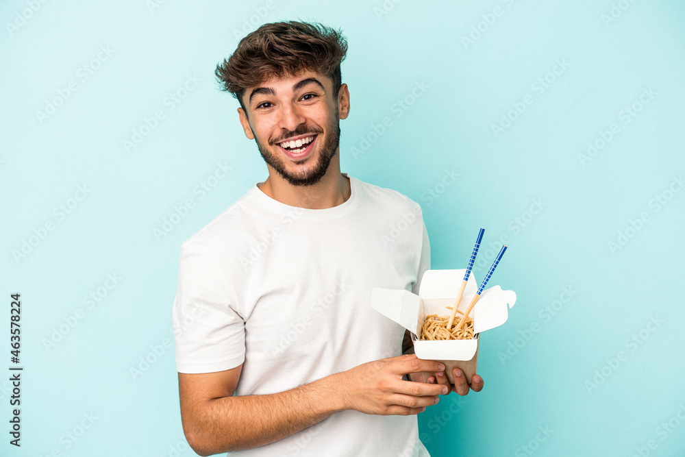 Young arab man holding a take away noodles isolated on blue background laughing and having fun. Young arab man holding a take away noodles isolated on blue background laughing and having fun.