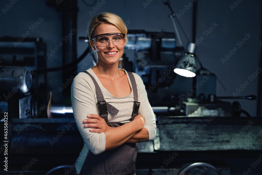 Portrait of mid adult industrial woman working indoors in metal workshop, looking at camera. - obrazy, fototapety, plakaty 