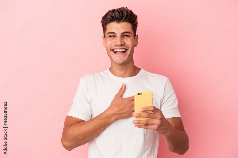 Young caucasian man holding a mobile phone isolated on pink background laughs out loudly keeping hand on chest. Young caucasian man holding a mobile phone isolated on pink background laughs out loudly keeping hand on chest.