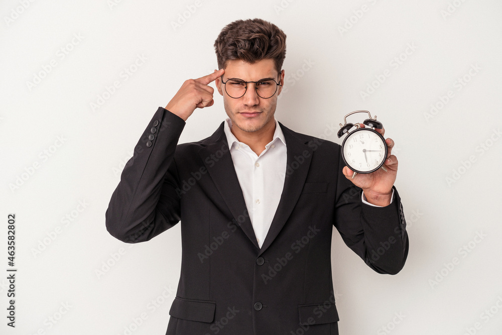 Young business caucasian man holding alarm clock isolated on white background pointing temple with finger, thinking, focused on a task. Young business caucasian man holding alarm clock isolated on white background pointing temple with finger, thinking, focused on a task.