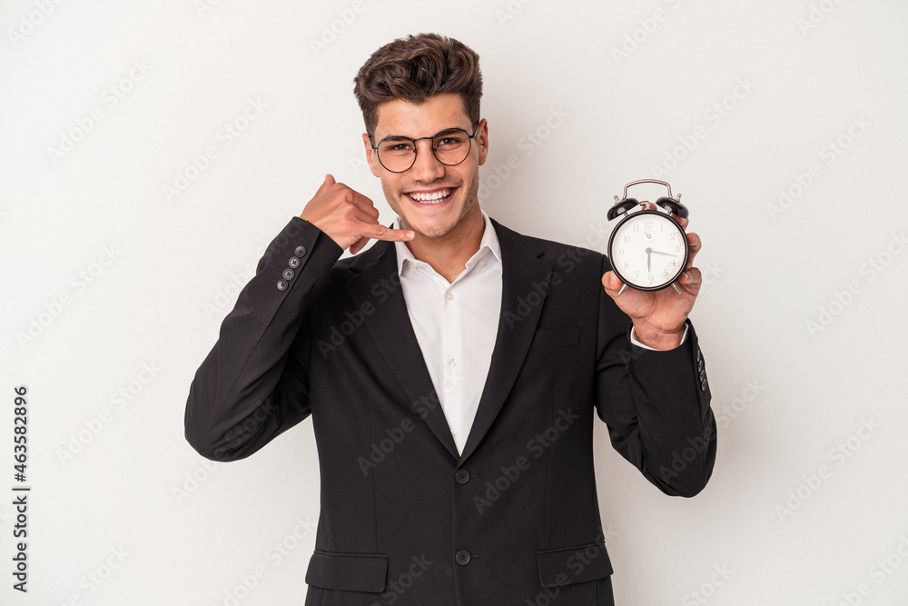 Young business caucasian man holding alarm clock isolated on white background showing a mobile phone call gesture with fingers. Young business caucasian man holding alarm clock isolated on white background showing a mobile phone call gesture with fingers.