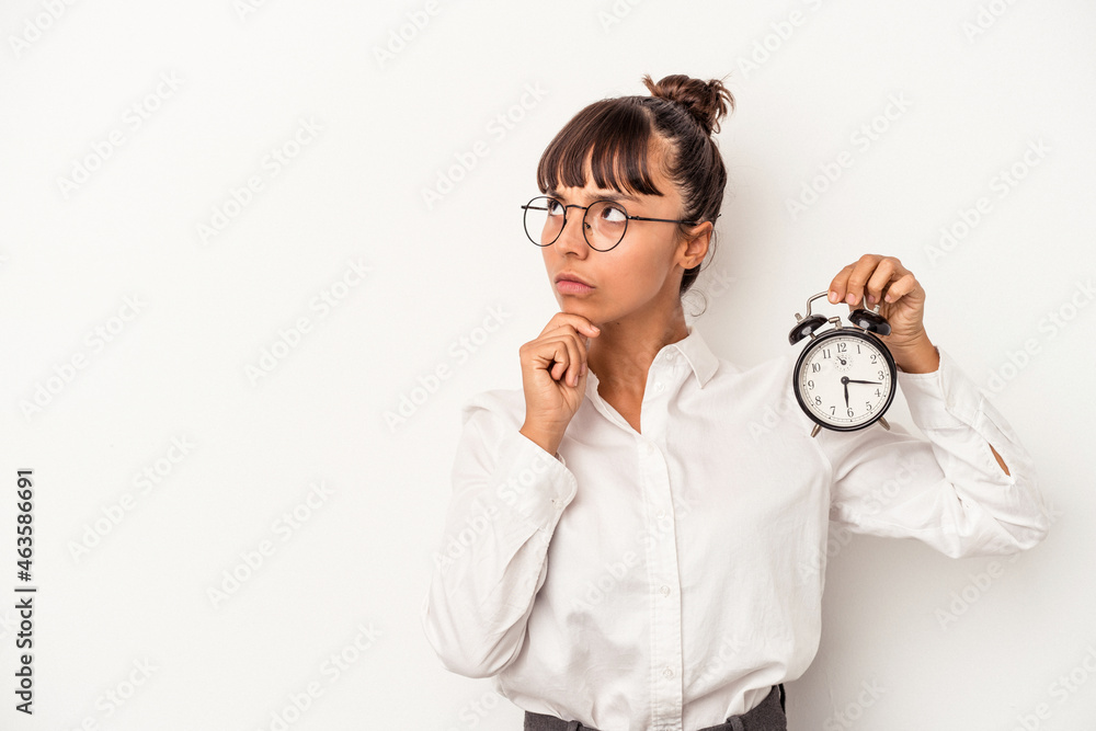Young mixed race business woman holding an alarm clock isolated on white background  looking sideways with doubtful and skeptical expression.