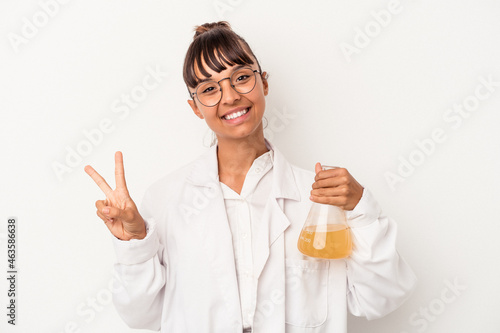 Young mixed race scientist woman holding a test tube isolated on white background  joyful and carefree showing a peace symbol with fingers.