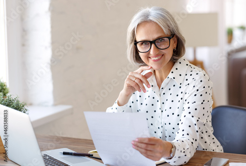 Smiling senior woman holding paper financial document and looking at laptop while working from home