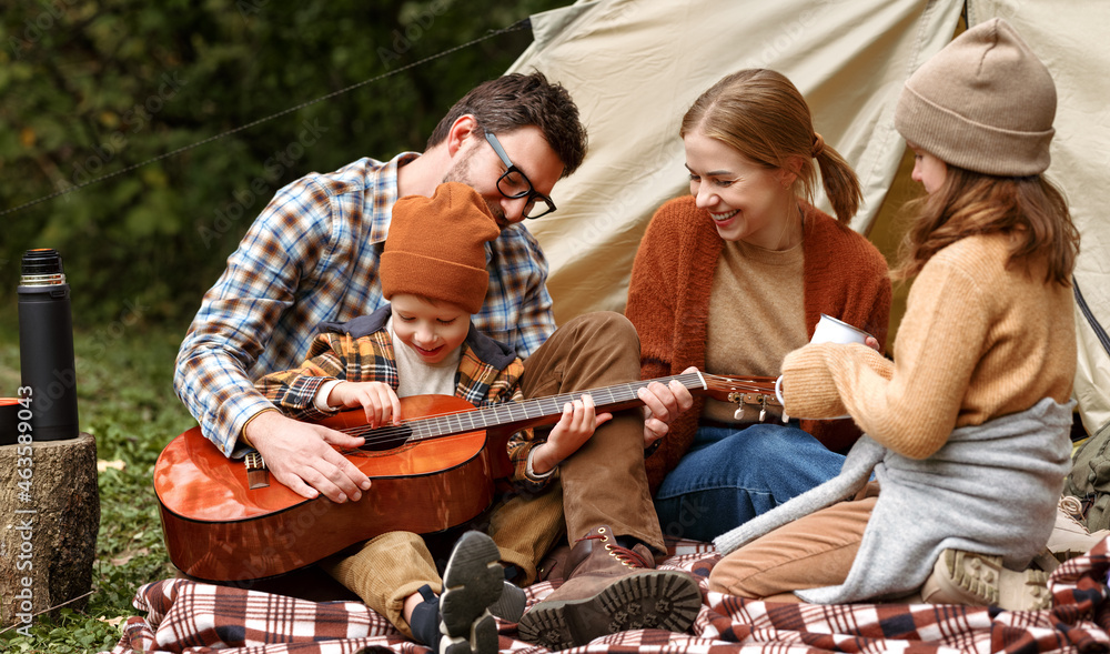 Father, mother and little son sitting near tourist tent and playing guitar during camping - obrazy, fototapety, plakaty 