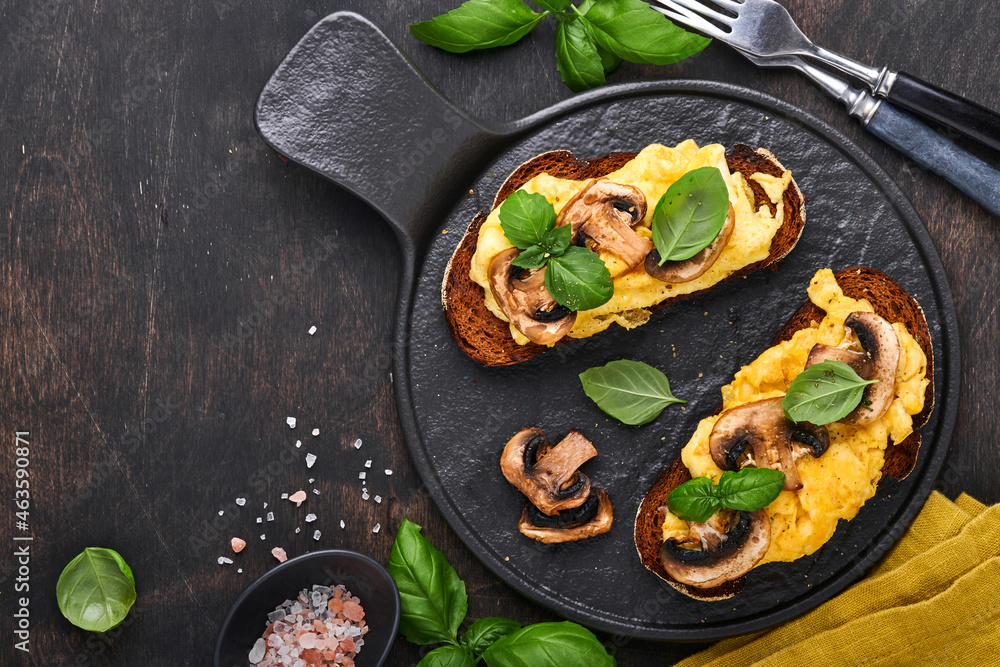 Scrambled eggs with fried mushrooms and basil on bread on black table background. Homemade breakfast or brunch meal - scrambled eggs and mushrooms sandwiches. Top view with copy space Scrambled eggs with fried mushrooms and basil on bread on black table background. Homemade breakfast or brunch meal - scrambled eggs and mushrooms sandwiches. Top view with copy space