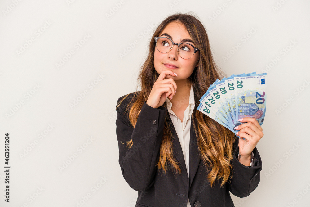 Young caucasian business woman holding banknotes isolated on white background looking sideways with doubtful and skeptical expression. Young caucasian business woman holding banknotes isolated on white background looking sideways with doubtful and skeptical expression.