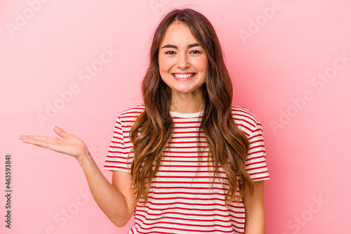 Young caucasian woman isolated on pink background showing a copy space on a palm and holding another hand on waist. © Asier