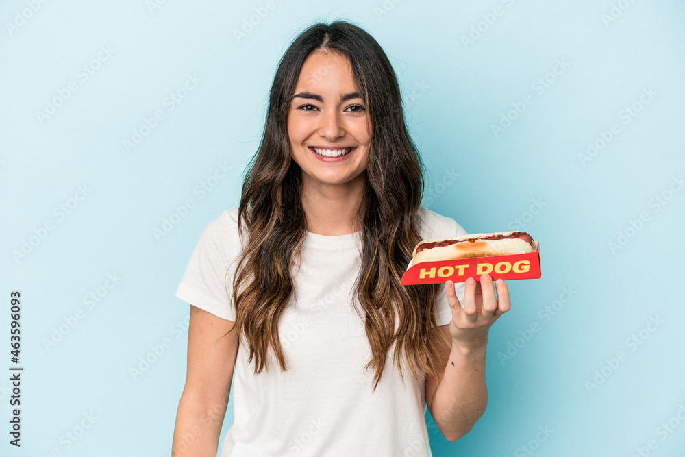 Young caucasian woman holding a hot dog isolated on blue background happy, smiling and cheerful. Young caucasian woman holding a hot dog isolated on blue background happy, smiling and cheerful.