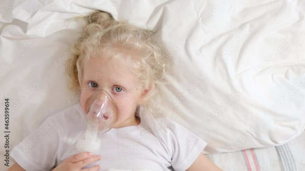cute little girl with curly blond hair lies in bed, sick five-tape girl in bed makes inhalation with a nebulizer, treatment of bronchitis and lung disease in children cute little girl with curly blond hair lies in bed, sick five-tape girl in bed makes inhalation with a nebulizer, treatment of bronchitis and lung disease in children