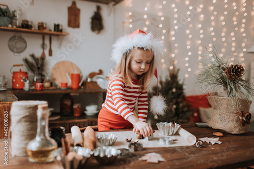 little girl in a Santa hat and red striped pajamas cooks a Christmas cake in a beautiful kitchen decorated with a garland. merry Christmas and happy New Year and Holidays. hygge. High quality photo