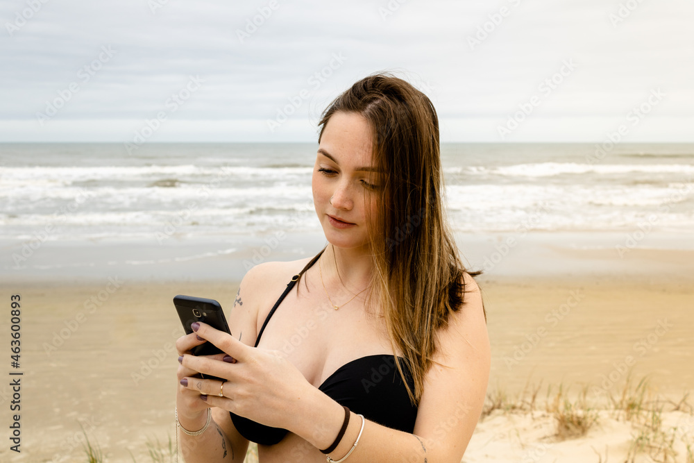 Girl in a bikini on the beach by the sea holding a cell phone sending a message on a cloudy day. Mention of vacations, travels, friendship, relationships and people. Contact with nature.