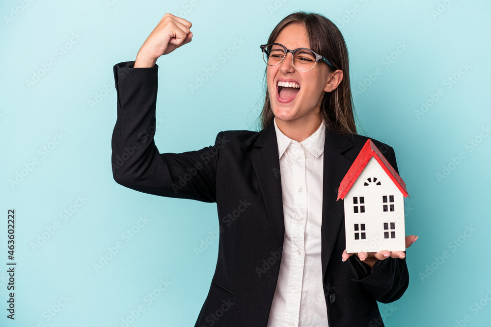 Young business woman holding a toy home isolated on blue background raising fist after a victory, winner concept. Young business woman holding a toy home isolated on blue background raising fist after a victory, winner concept.