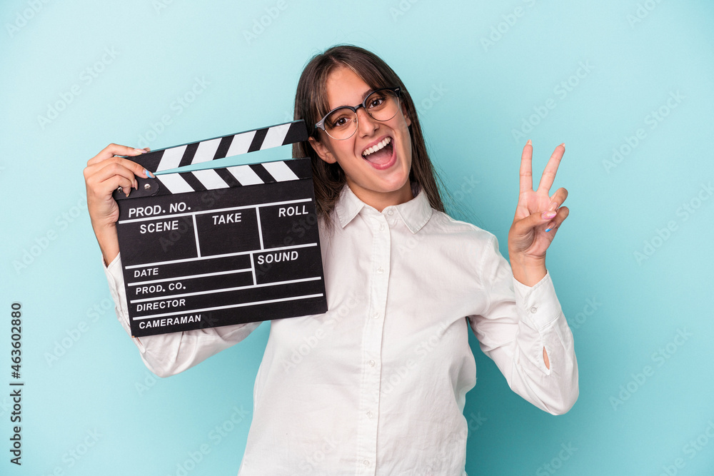 Young caucasian woman holding clapperboard isolated on blue background joyful and carefree showing a peace symbol with fingers. Young caucasian woman holding clapperboard isolated on blue background joyful and carefree showing a peace symbol with fingers.