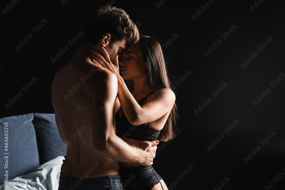 Naklejka premium Pretty woman in lingerie kissing shirtless boyfriend on blurred bed isolated on black
