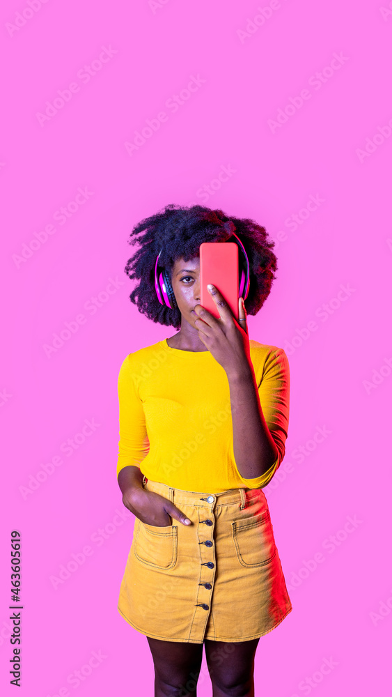 Young black woman covering her face with smartphone wearing wireless technology headphones isolated advertising copy space background