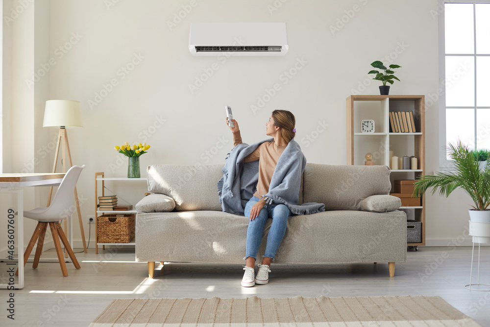 Woman who's sitting on sofa under warm plaid in living room switches off her air conditioner on wall. Young girl adjusting modern AC system, regulating temperature and enjoying cool fresh air at home - obrazy, fototapety, plakaty 