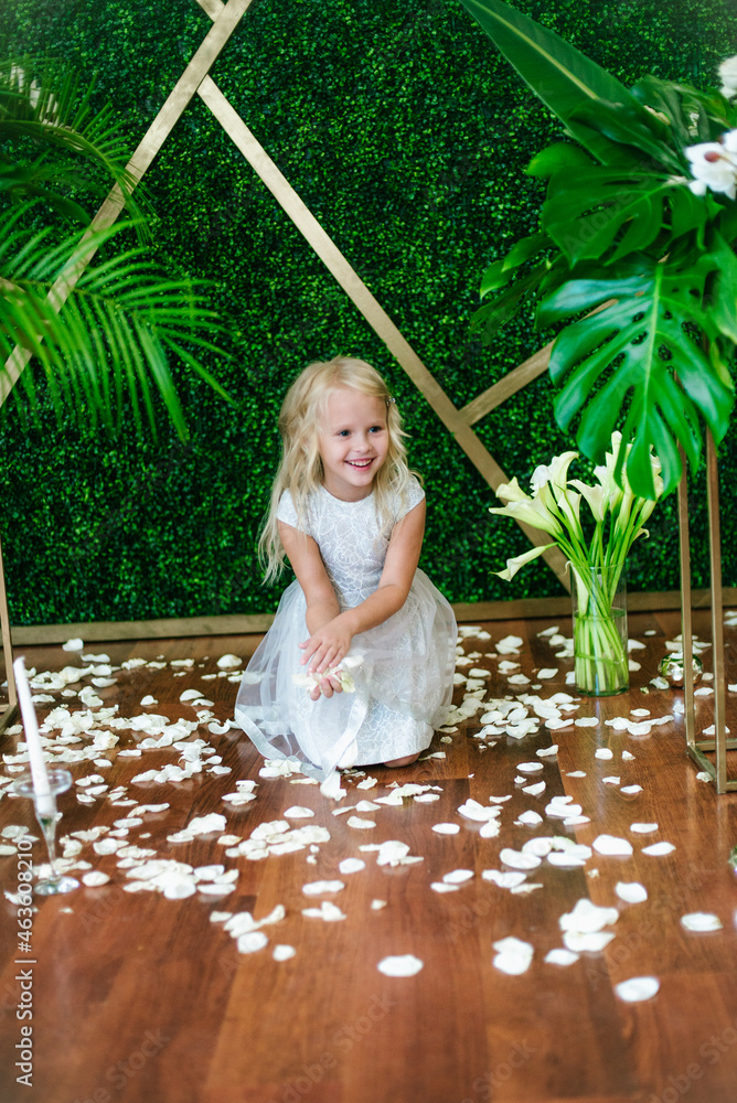 Little cute girl with blond hair in a white dress and white flowers, lilies and orchids on a background with a green tropical plants Little cute girl with blond hair in a white dress and white flowers, lilies and orchids on a background with a green tropical plants