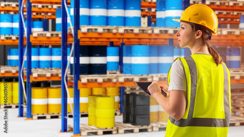 manager is counting chemical barrels. Metal barrels in front of warehouse worker. Manager in yellow uniform. Blurred racks in background. Work in chemical industry. Warehouse worker with tablet photo