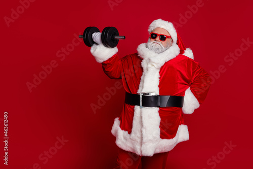 Portrait of attractive bearded funny Santa lifting dumbbell working out difficult hard isolated over bright red color background