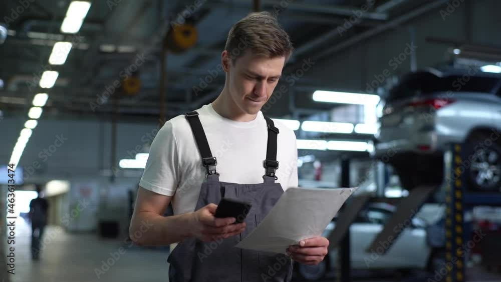 Portrait of serious handsome mechanic male in blue uniform dialing customer number for call. Auto technician text messaging on cell phone in workshop holding report. Tracking shot in slow motion. Portrait of serious handsome mechanic male in blue uniform dialing customer number for call. Auto technician text messaging on cell phone in workshop holding report. Tracking shot in slow motion.
