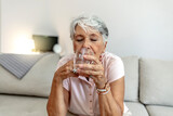 Senior woman taking tablet with glass of water at home. Mature woman taking pill against headaches at home. Woman taking medication for her illness. Gray hair woman taking pill to ease headache