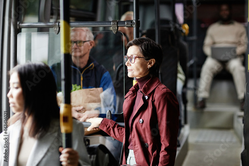 Side view portrait of adult woman holding onto railing in bus while traveling by public transport in city, copy space