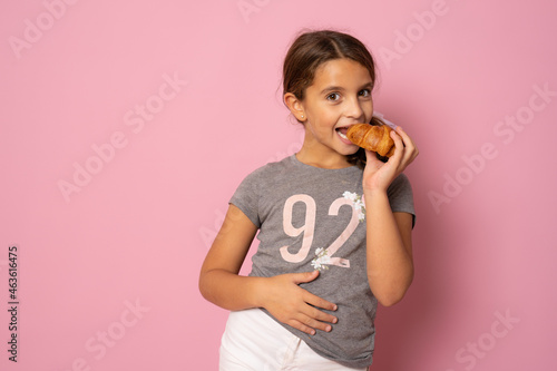 Smiling happy little girl eating croissant with pleasure. Studio shot isolated over pink background.