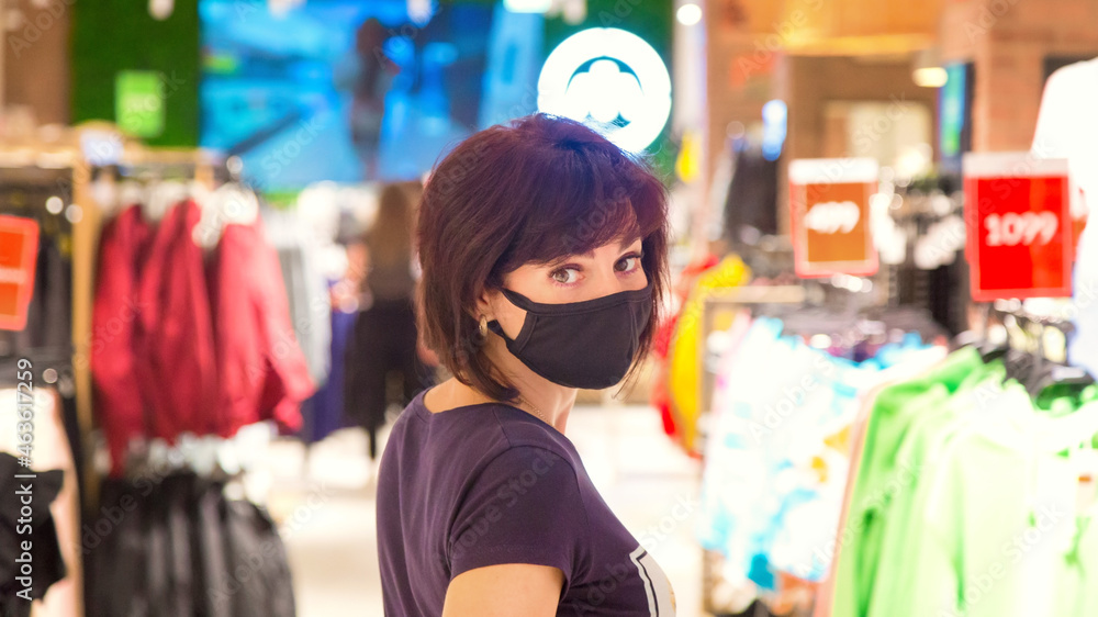 woman in a black protective mask in shopping mall among hangers with things during the pandemic. woman in a black protective mask in shopping mall among hangers with things during the pandemic.