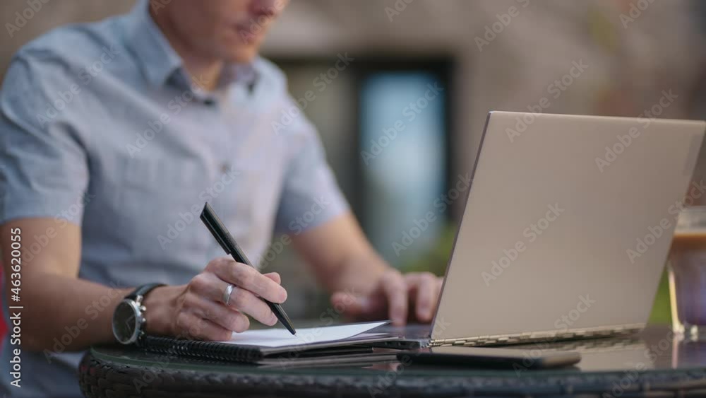 A man sitting in a street cafe works remotely writing a pen in a notebook. Work at the laptop. A man works remotely on a laptop A man sitting in a street cafe works remotely writing a pen in a notebook. Work at the laptop. A man works remotely on a laptop